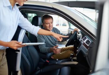A man sitting in a car being instructed on how to use the vehicle by a woman with a tablet